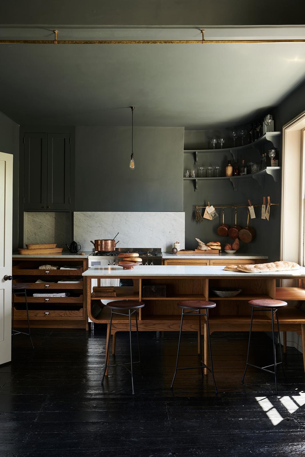 A cosy and sophisticated Haberdasher’s kitchen features a cooker run with a Carrara marble worktop, a small prep table, bespoke curved shelves, and dark charcoal walls. Beautifully decorated with stools, aged brass hanging rails, copper pans, fresh bread, vintage items, and artisan ceramics, this kitchen creates a cool yet warm and inviting space.