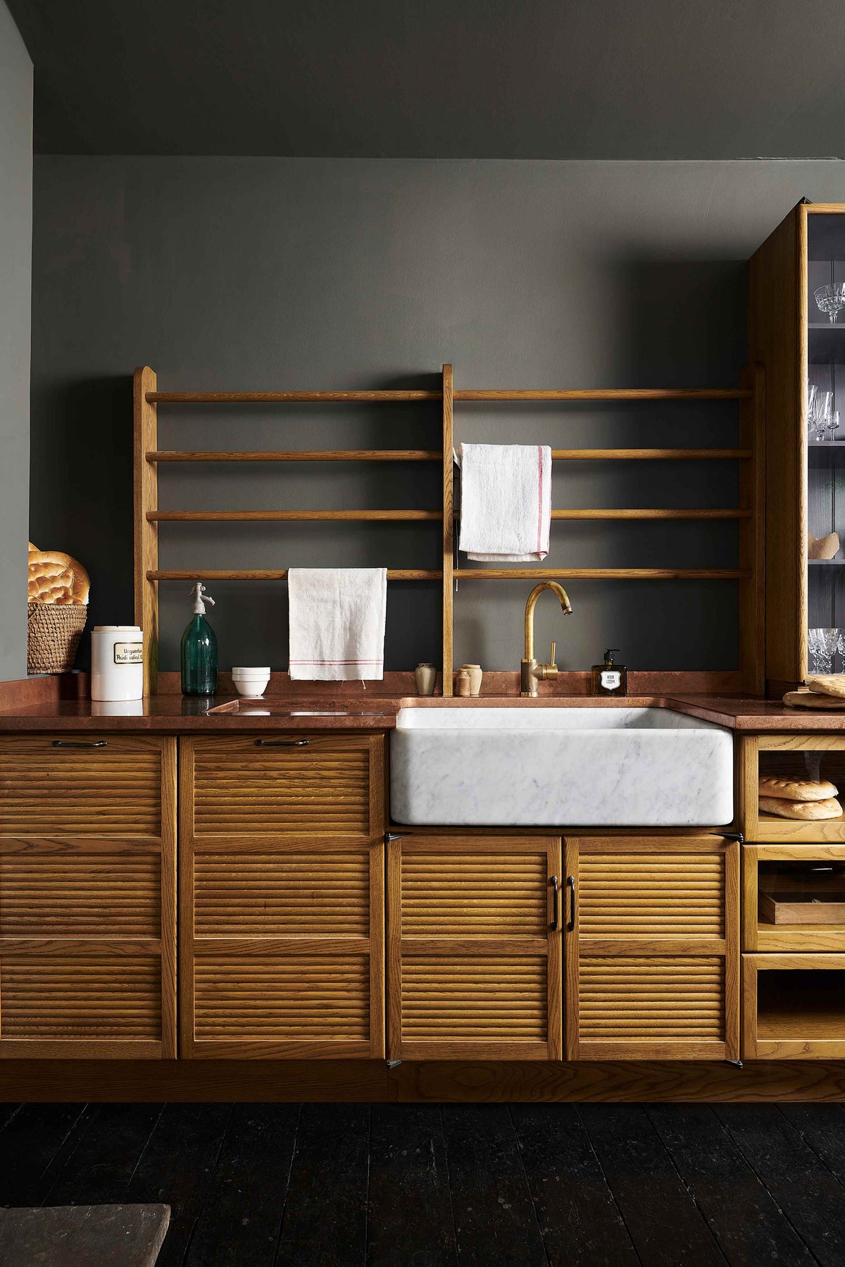 A closer look at a cosy and sophisticated Haberdasher’s kitchen features a sink run with a copper worktop, a single marble sink, glazed countertop cupboards, a freestanding pantry with glazed doors, and dark charcoal walls. Beautifully decorated with vintage furniture and items, wicker baskets, fresh bread, tea towels, and artisan ceramics, this kitchen creates a bright and warm space.