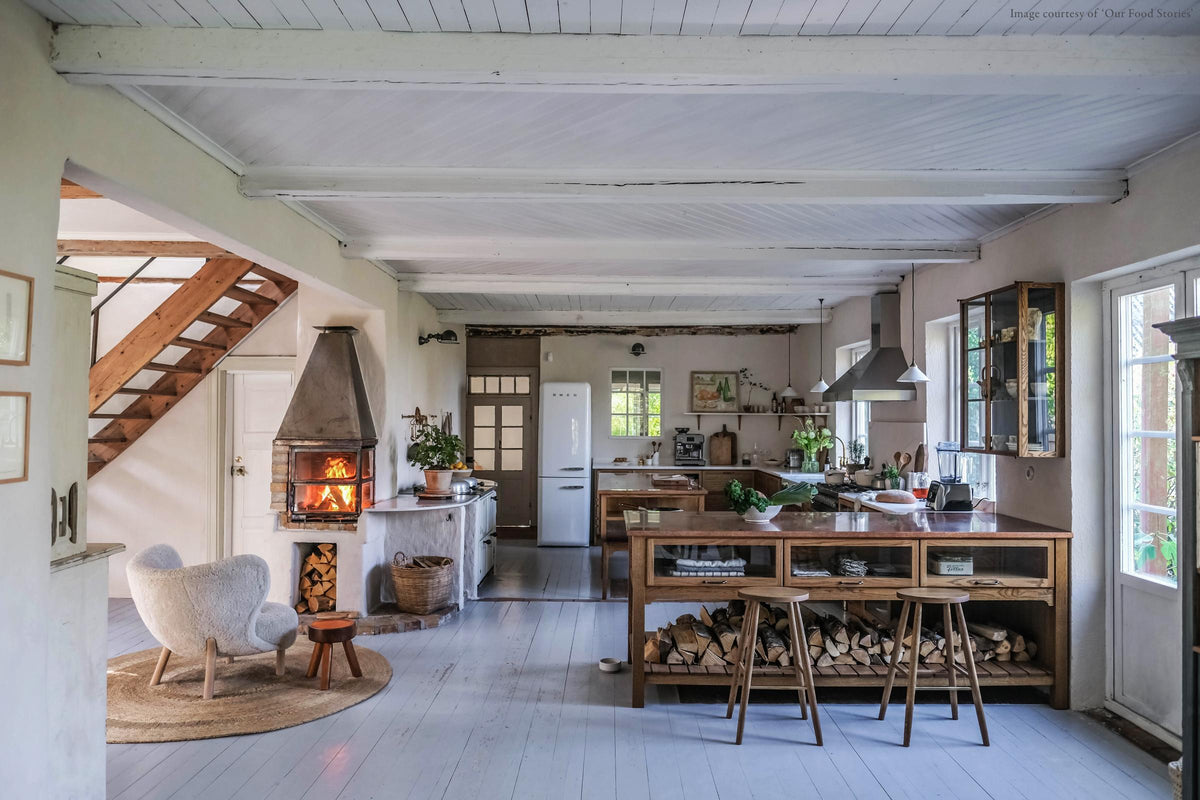 A warm and inviting Haberdasher’s kitchen features a sink and cooker run with a Carrara marble worktop, a double-fluted marble sink, an aged brass Ionian tap, prep tables with a copper worktop, glazed wall cupboards, and a shelf. Beautifully decorated with vintage furniture and items, artisan ceramics, firewood, a small fireplace, and a wool armchair, this kitchen creates an airy and cosy Scandinavian small fishing town hideaway.