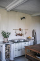 An oblique view of a warm and inviting Haberdasher’s kitchen features a cooker run with a Carrara marble worktop and prep tables with a copper worktop. Beautifully decorated with vintage furniture and items, artisan ceramics, firewood, aged brass hanging rails, and a white gas stove, this kitchen creates an airy and cosy Scandinavian small fishing town hideaway.