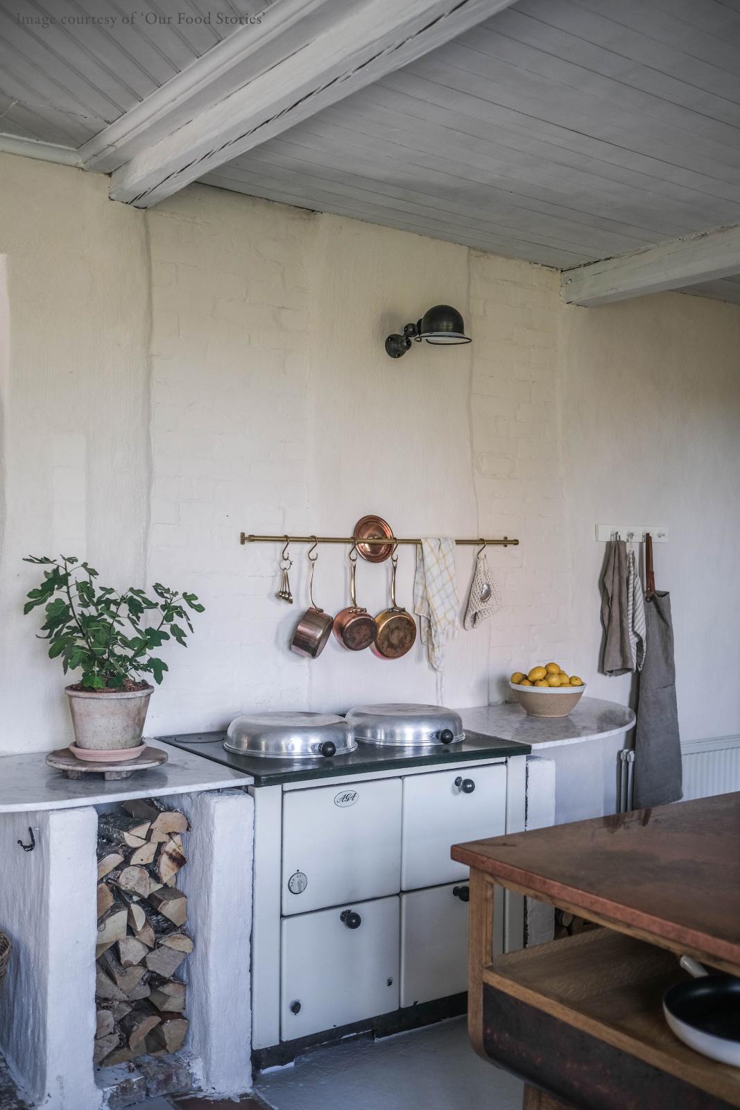 An oblique view of a warm and inviting Haberdasher’s kitchen features a cooker run with a Carrara marble worktop and prep tables with a copper worktop. Beautifully decorated with vintage furniture and items, artisan ceramics, firewood, aged brass hanging rails, and a white gas stove, this kitchen creates an airy and cosy Scandinavian small fishing town hideaway.