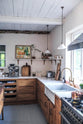 An oblique view of a warm and inviting Haberdasher’s kitchen features a sink run with a Carrara marble worktop, a double-fluted marble sink, an aged brass Ionian tap, a shelf, and prep tables with a copper worktop. Beautifully decorated with vintage furniture and items, artisan ceramics, a still life painting, and classic shelf brackets, this kitchen creates an airy and cosy Scandinavian small fishing town hideaway.