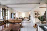 A warm and inviting Haberdasher’s kitchen features a sink and cooker run with a Carrara marble worktop, a double-fluted marble sink, an aged brass Ionian tap, prep tables with a copper worktop, and glazed wall cupboards. Beautifully decorated with vintage furniture and items, artisan ceramics, firewood, a small fireplace, and a wool armchair, this kitchen creates an airy and cosy Scandinavian small fishing town hideaway.