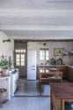 A warm and inviting Haberdasher’s kitchen features a sink and cooker run with a Carrara marble worktop, a double-fluted marble sink, an aged brass Ionian tap, prep tables with a copper worktop, and a shelf. Beautifully decorated with vintage furniture and items, artisan ceramics, a wicker basket, and a white refrigerator, this kitchen creates an airy and cosy Scandinavian small fishing town hideaway.