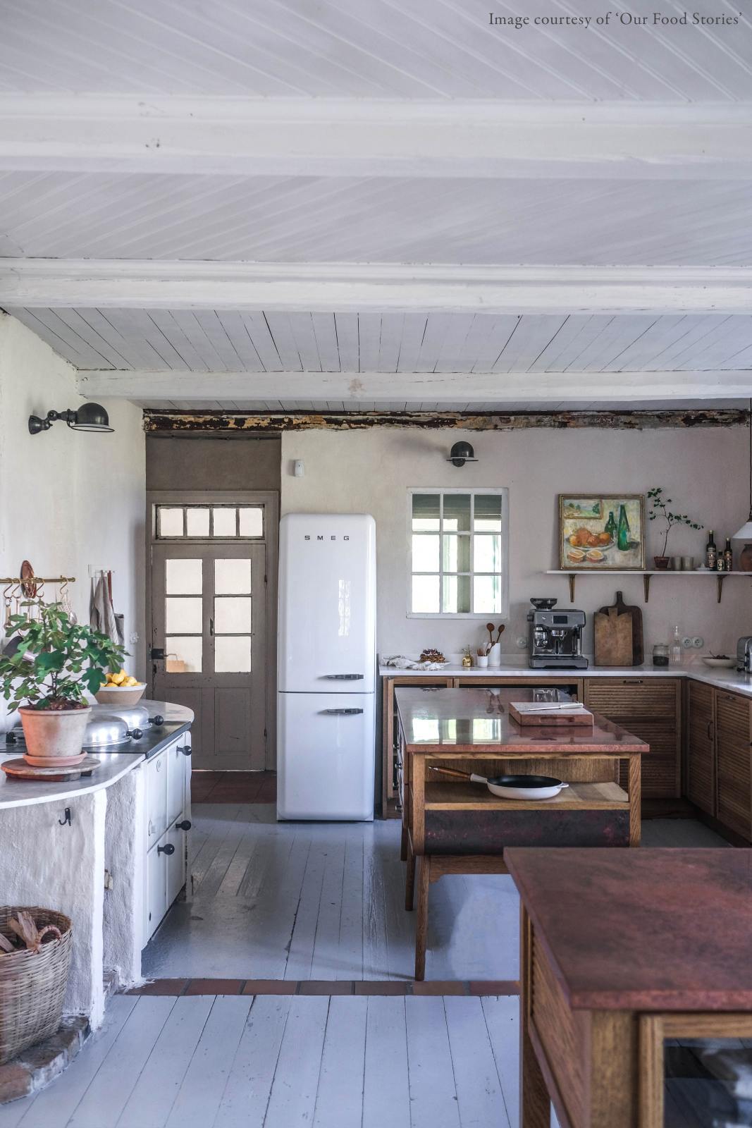 A warm and inviting Haberdasher’s kitchen features a sink and cooker run with a Carrara marble worktop, a double-fluted marble sink, an aged brass Ionian tap, prep tables with a copper worktop, and a shelf. Beautifully decorated with vintage furniture and items, artisan ceramics, a wicker basket, and a white refrigerator, this kitchen creates an airy and cosy Scandinavian small fishing town hideaway.