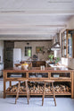 A warm and inviting Haberdasher’s kitchen features a sink and cooker run with a Carrara marble worktop, a double-fluted marble sink, an aged brass Ionian tap, prep tables with a copper worktop, glazed wall cupboards, and a shelf. Beautifully decorated with vintage furniture and items, artisan ceramics, firewood, Helen stools, and a bowl of lemons, this kitchen creates an airy and cosy Scandinavian small fishing town hideaway.