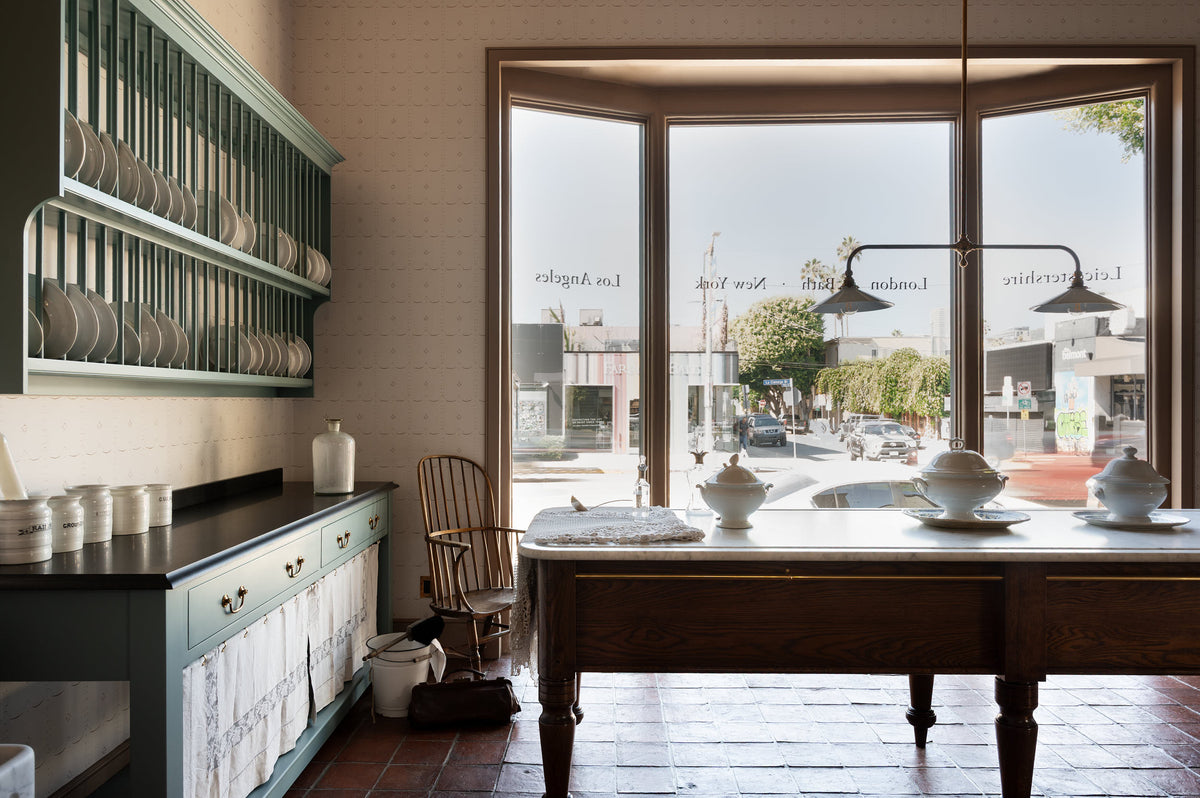 Looking out towards the large window of a beautiful and functional Trinity blue Heirloom kitchen features open cabinets, plate racks, and a showpiece Dairy table. Lovingly adorned with bone-white artisan ceramics, selected antiques, lace curtains, Gaselier lights, and earthy red terracotta square floor tiles that recreate Victorian kitchen furniture made to last a lifetime.