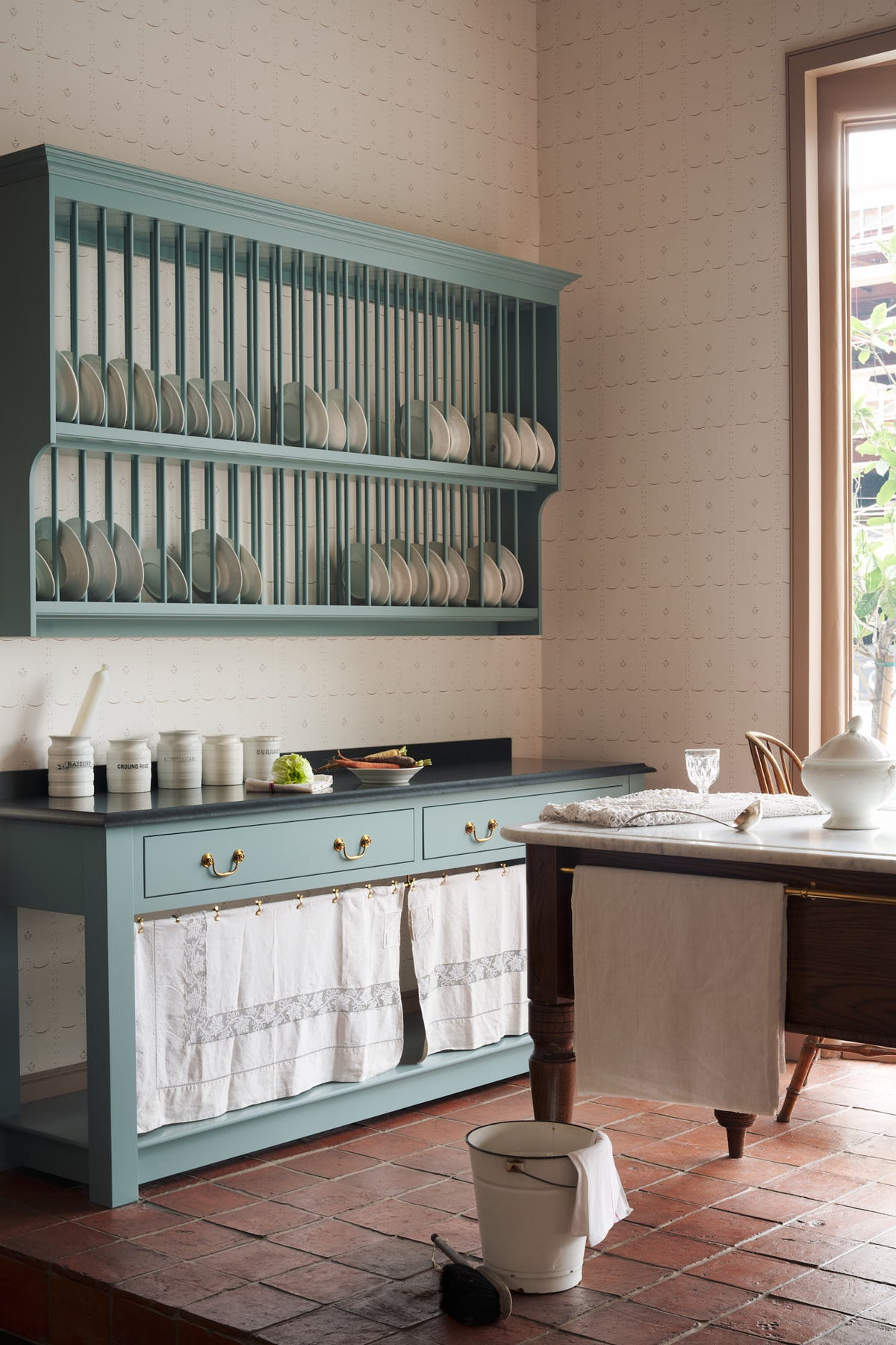 An oblique view of a beautiful and functional Trinity blue Heirloom kitchen features open cabinets, plate racks, and a showpiece Dairy table. Lovingly adorned with bone-white artisan ceramics, selected antiques, lace curtains, Gaselier lights, and earthy red terracotta square floor tiles that recreate Victorian kitchen furniture made to last a lifetime.