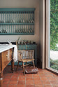A quiet corner of a beautiful and functional Trinity blue Heirloom kitchen features open cabinets, plate racks, and a showpiece Dairy table. Lovingly adorned with bone-white artisan ceramics, selected antiques, lace curtains, and earthy red terracotta square floor tiles that recreate Victorian kitchen furniture made to last a lifetime.