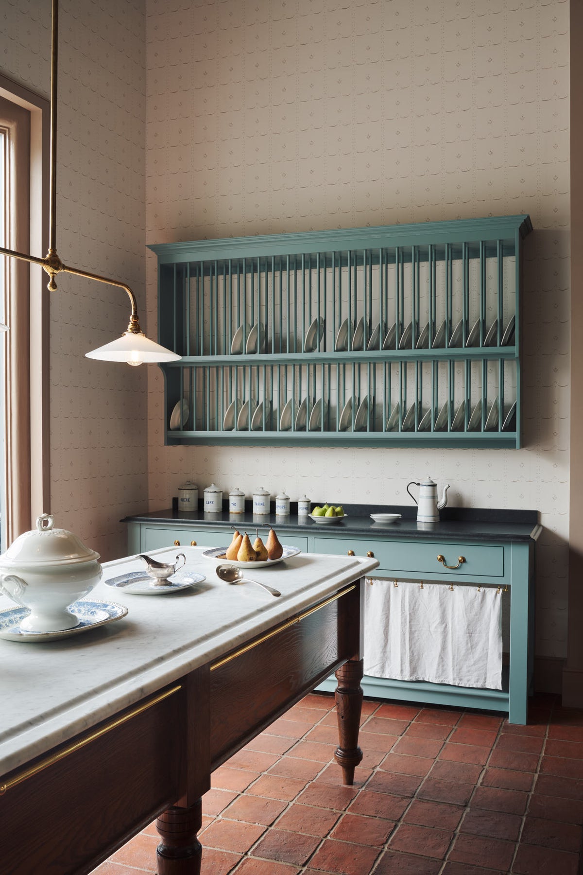 An oblique view through a doorway of a beautiful and functional Trinity blue Heirloom kitchen features open cabinets, plate racks, and a showpiece Dairy table. Lovingly adorned with bone-white artisan ceramics, selected antiques, lace curtains, Gaselier lights, and earthy red terracotta square floor tiles that recreate a Victorian kitchen made to last a lifetime.