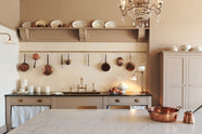 A grand Princelet pink Heirloom and Classic English kitchen features a sink cabinet with a black granite worktop, a butler’s pantry, a large kitchen island with a Carrara marble worktop, aged brass rails, ditsy delft wallpaper, a tall cream subway tile splashback, a grand tour desk lamp, and an Heirloom task light. Lovingly adorned with brass pots and pans, artisan ceramics, and an antique crystal chandelier, creating a warm, delicate, and airy kitchen space.
