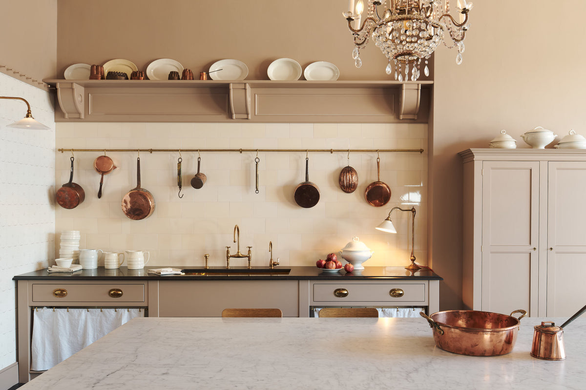 A grand Princelet pink Heirloom and Classic English kitchen features a sink cabinet with a black granite worktop, a butler’s pantry, a large kitchen island with a Carrara marble worktop, aged brass rails, ditsy delft wallpaper, a tall cream subway tile splashback, a grand tour desk lamp, and an Heirloom task light. Lovingly adorned with brass pots and pans, artisan ceramics, and an antique crystal chandelier, creating a warm, delicate, and airy kitchen space.