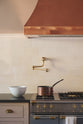 A close-up of an aged brass pot filler tap against tall cream subway tile splashback, and a large copper canopy. Adorned with a brass pan, a white ceramic bowl and a tea towel.