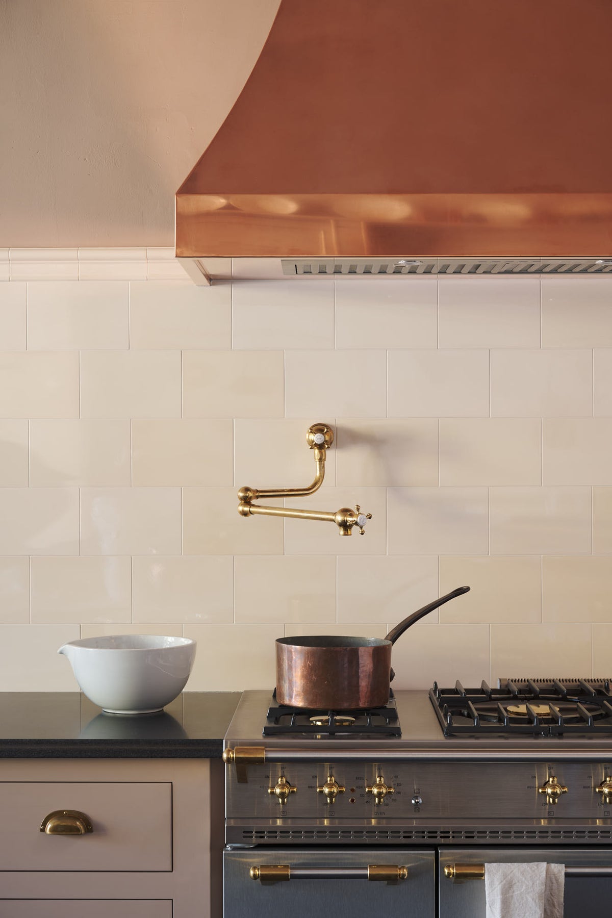 A close-up of an aged brass pot filler tap against tall cream subway tile splashback, and a large copper canopy. Adorned with a brass pan, a white ceramic bowl and a tea towel.