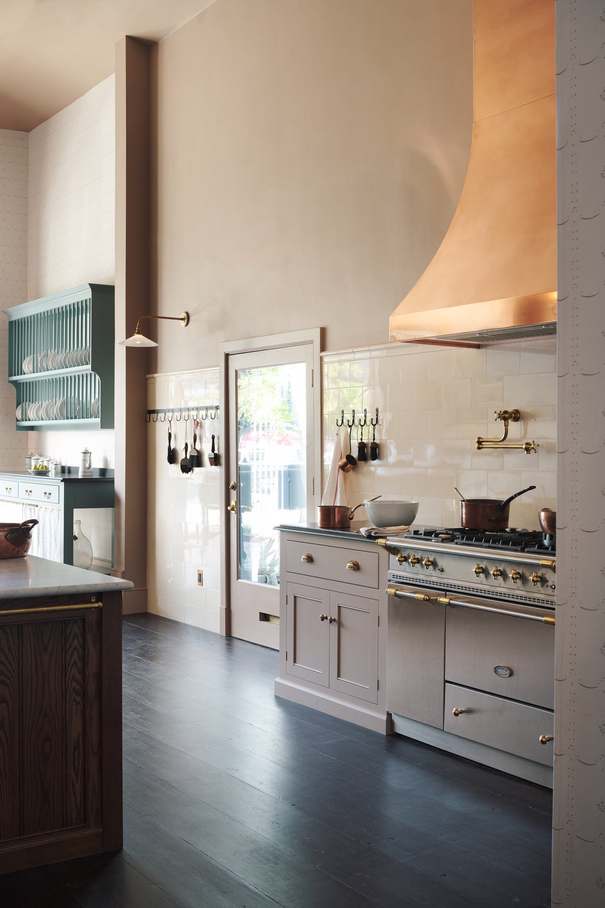 An oblique view of a quiet corner of a grand Princelet Pink Heirloom and Classic English kitchen features a cooker run with a black granite worktop, a large kitchen island with a Carrara marble worktop, an aged brass pot filler tap against a tall cream subway tile and border tile splashback, and a large copper canopy. Lovingly adorned with brass pots and pans, and artisan ceramics, creating a warm, delicate, and airy kitchen space.
