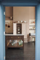 Looking through the doorway, you see a grand Princelet Pink Heirloom and Classic English kitchen. It features a Butler’s pantry cupboard, a large kitchen island with a Carrara marble worktop, and a janitor’s sink area with a tall cream subway and border-tile splashback. The space is lovingly adorned with brass pots and pans and artisan ceramics, creating a warm, delicate, and airy feel.