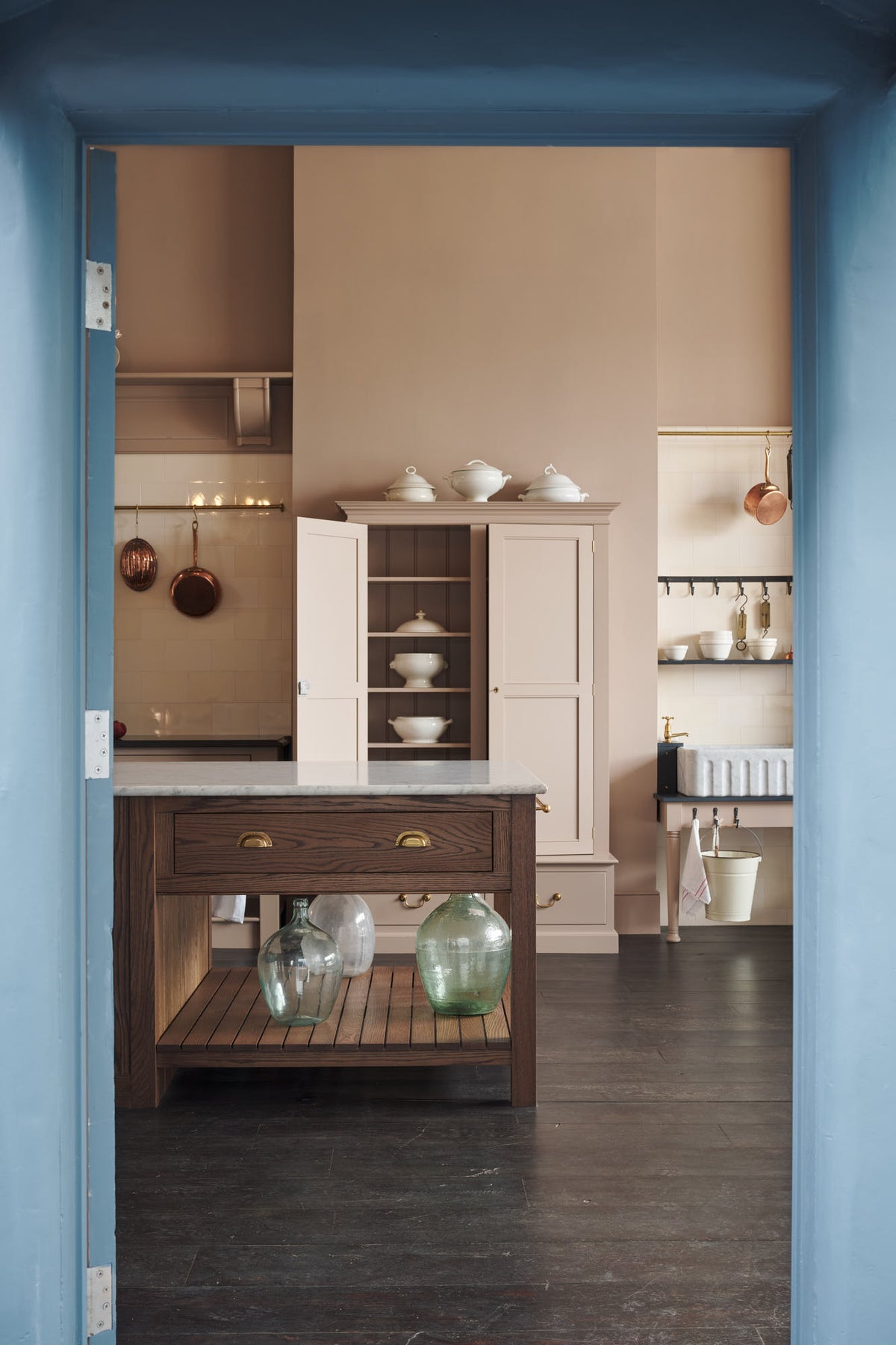 Looking through the doorway, you see a grand Princelet Pink Heirloom and Classic English kitchen. It features a Butler’s pantry cupboard, a large kitchen island with a Carrara marble worktop, and a janitor’s sink area with a tall cream subway and border-tile splashback. The space is lovingly adorned with brass pots and pans and artisan ceramics, creating a warm, delicate, and airy feel.