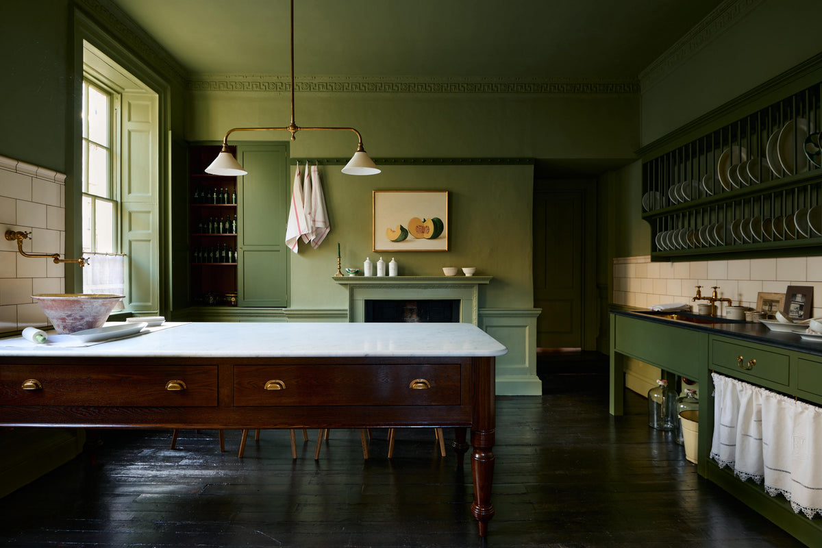 A quaint and stylish Wilkes green Heirloom kitchen features a sink cabinet with a café rail, white lace curtains, a polished brass sink, aged brass Mayan taps, a large plate rack, and a dairy table. Adorned with an aged brass pot filler, artisan ceramics, Heirloom gaselier lights, cream subway splashback tiles, and a golden-framed sliced green pumpkin, this kitchen creates a luxurious dining and social space.