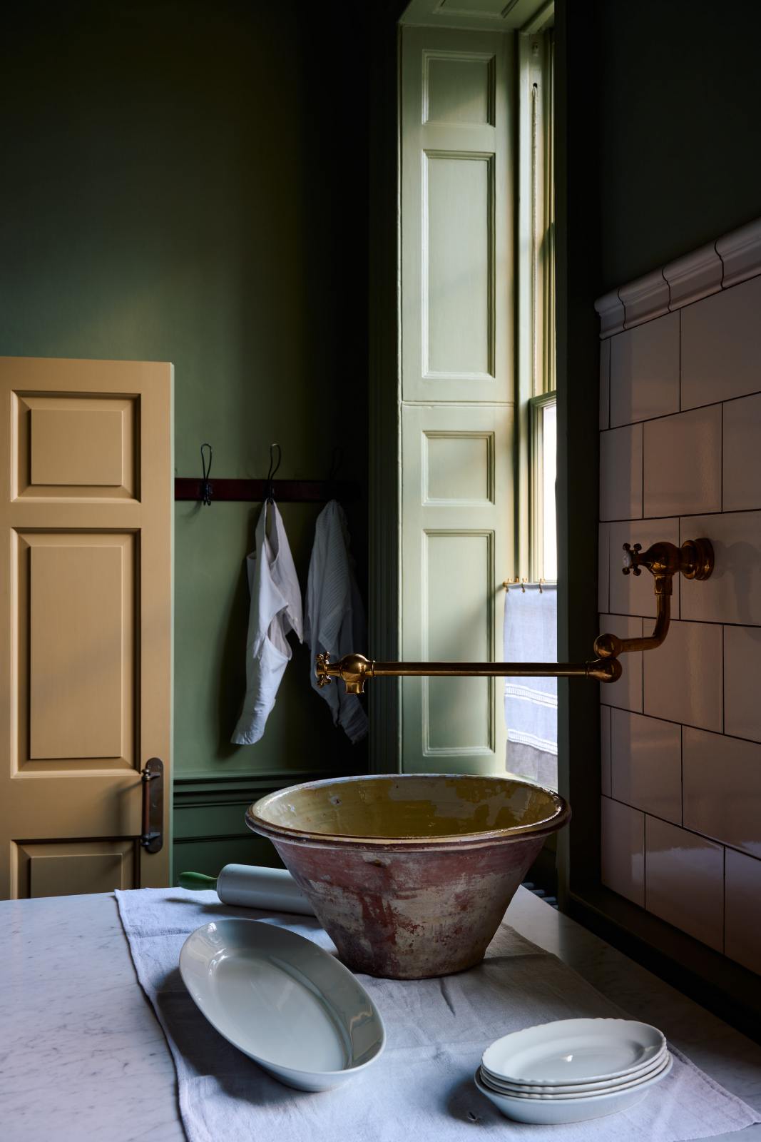 A close-up of a quaint and stylish Wilkes green Heirloom kitchen features a large dairy table. Adorned with an aged brass pot filler, artisan ceramics, and cream subway splashback tiles, this kitchen creates a luxurious dining and social space.