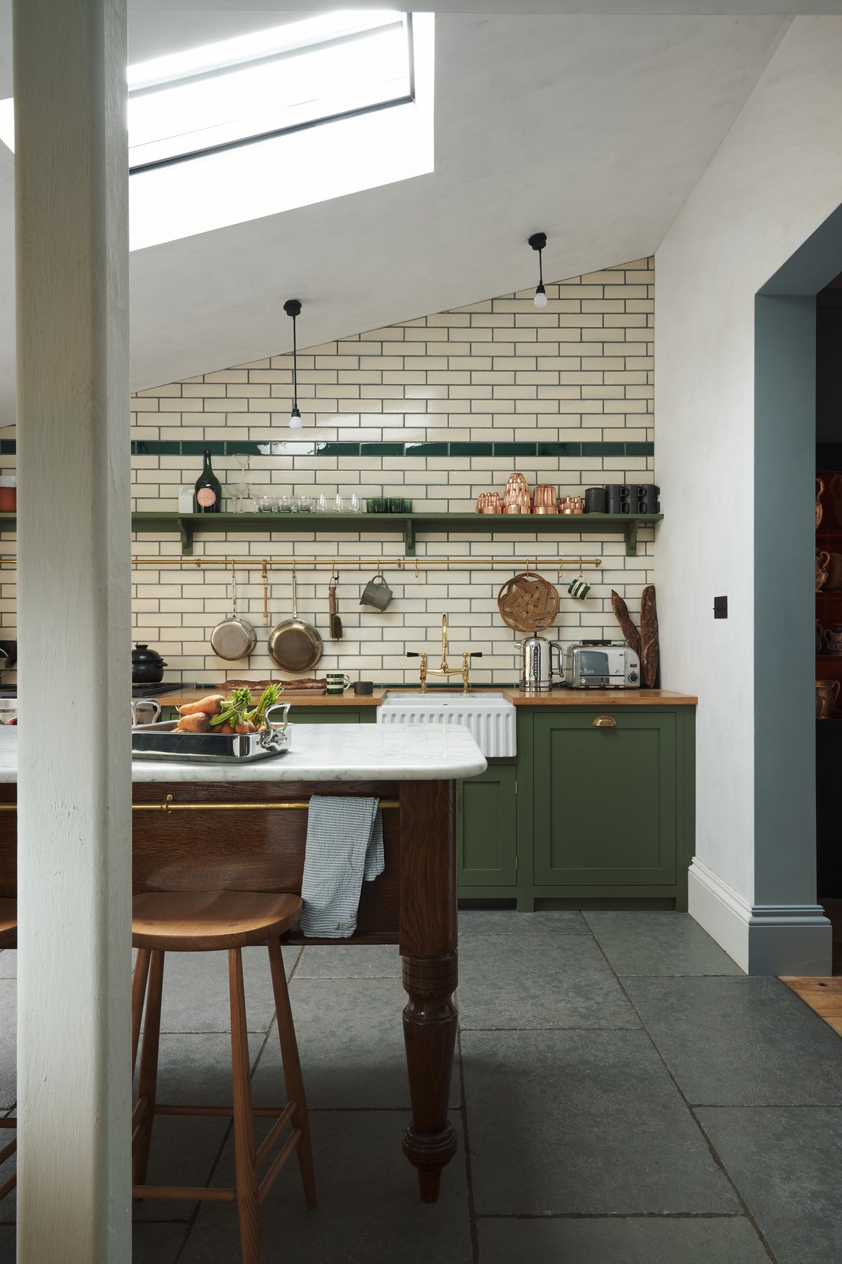 A quiet corner of a quaint and modern Wilkes green Shaker kitchen features a cooker and sink run with a rustic oak worktop, a long shaker shelf, and a Dairy table with a breakfast nook. Adorned with a long hanging rail, signature ceramics, brass cups, and a large wall of metro tiles it embodies a playful industrial and practical design. 