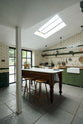 An oblique view of a quaint and modern Wilkes green Shaker kitchen features a cooker and sink run with a rustic oak worktop, a long shaker shelf, and a Dairy table with a breakfast nook. Adorned with a long hanging rail, sky light, signature ceramics, brass cups, and a large wall of metro tiles it embodies a playful industrial and practical design.