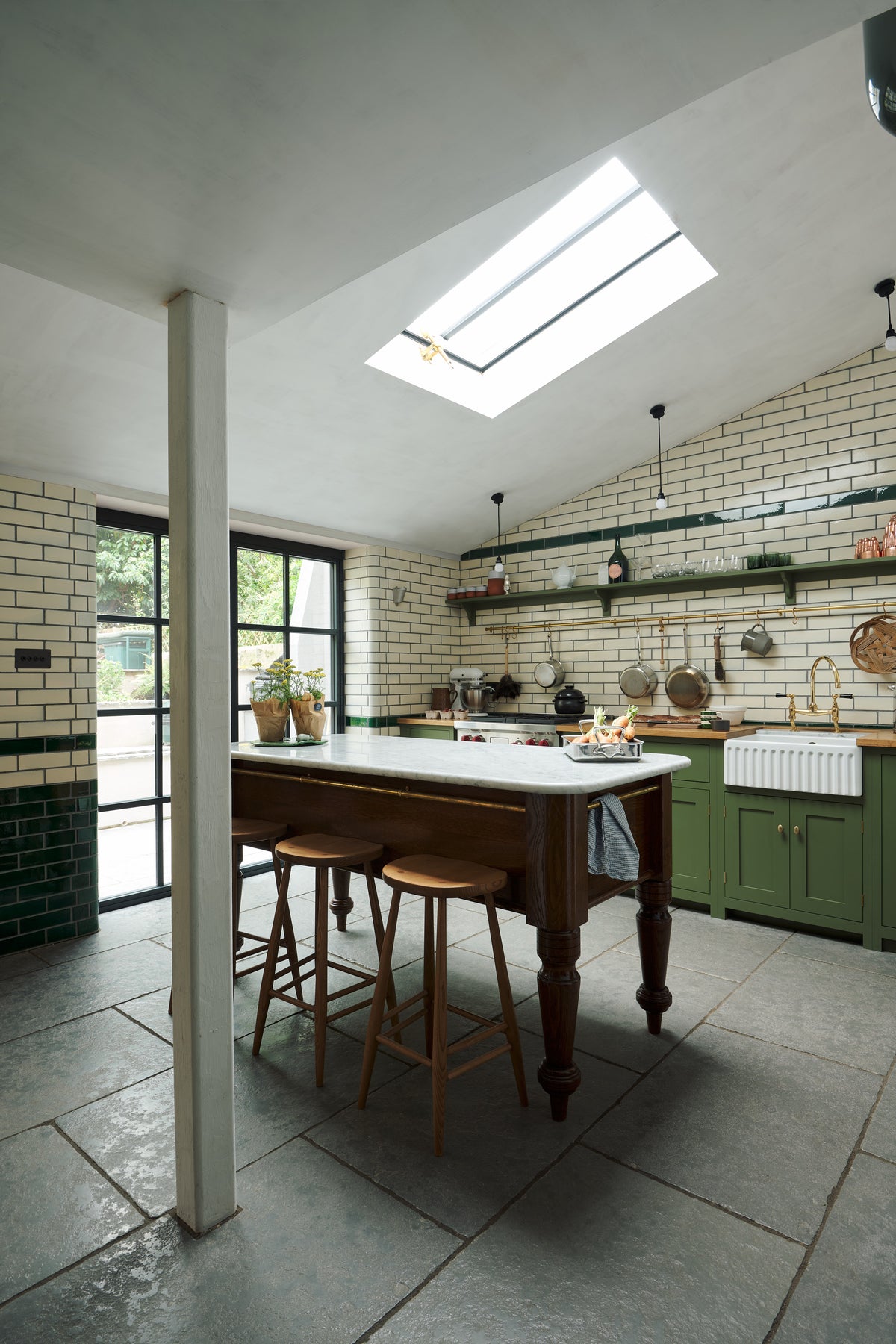 An oblique view of a quaint and modern Wilkes green Shaker kitchen features a cooker and sink run with a rustic oak worktop, a long shaker shelf, and a Dairy table with a breakfast nook. Adorned with a long hanging rail, sky light, signature ceramics, brass cups, and a large wall of metro tiles it embodies a playful industrial and practical design.