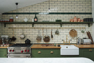 A closer look at the quaint and modern Wilkes green Shaker kitchen features a cooker and sink run with a rustic oak worktop, a long shaker shelf, and a Dairy table with a breakfast nook. Adorned with a long hanging rail, signature ceramics, brass cups, and a large wall of metro tiles it embodies a playful industrial and practical design.
