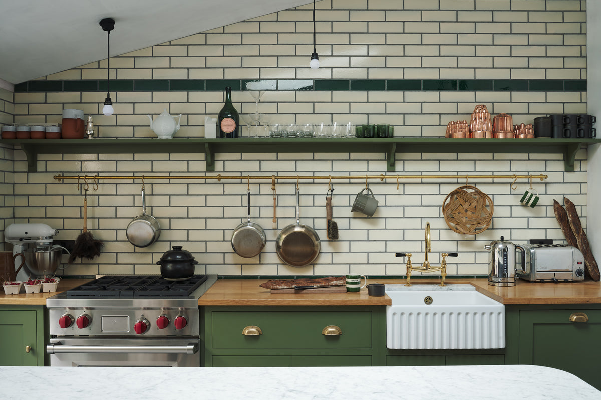 A closer look at the quaint and modern Wilkes green Shaker kitchen features a cooker and sink run with a rustic oak worktop, a long shaker shelf, and a Dairy table with a breakfast nook. Adorned with a long hanging rail, signature ceramics, brass cups, and a large wall of metro tiles it embodies a playful industrial and practical design.