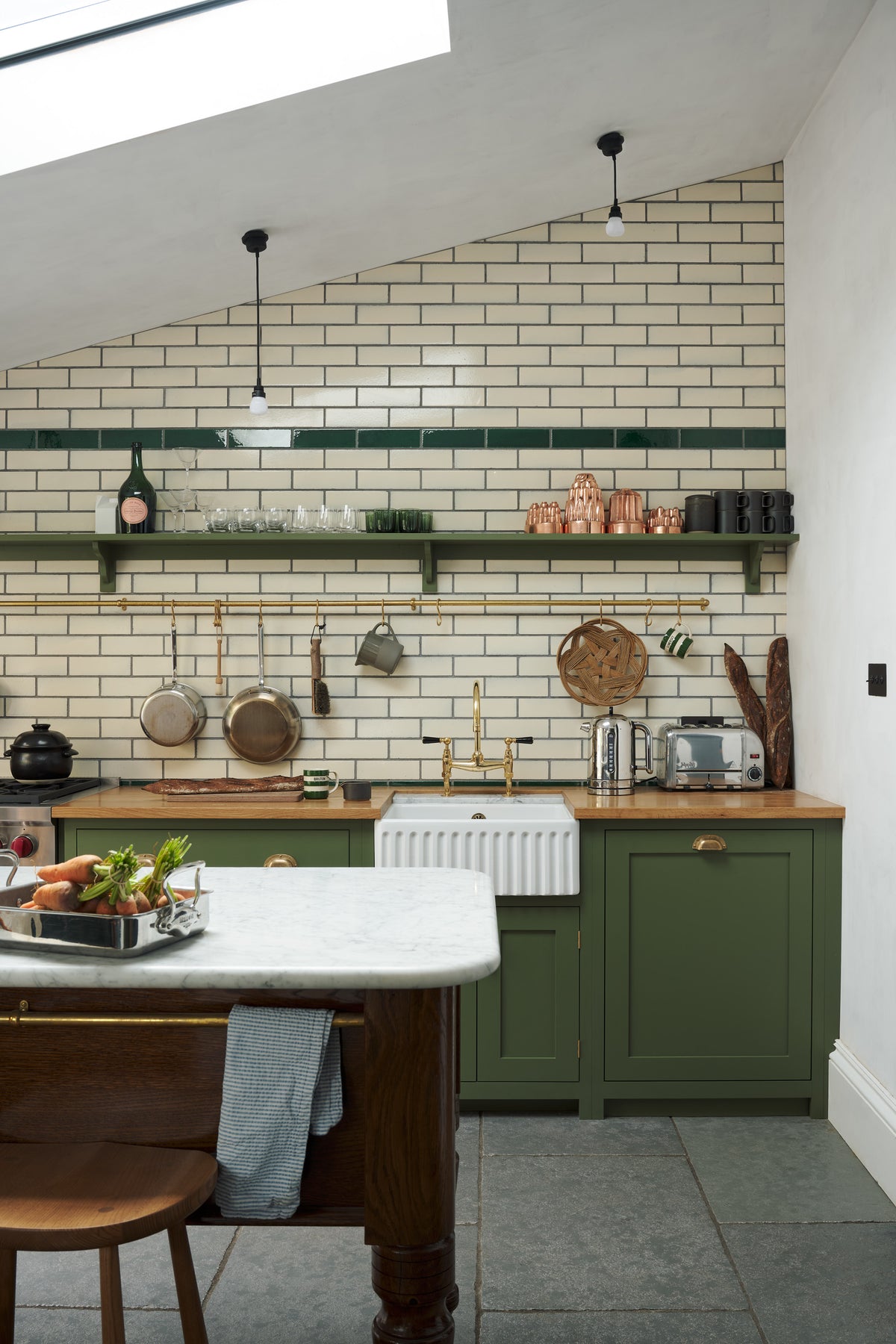 A quiet corner of a quaint and modern Wilkes green Shaker kitchen features a cooker and sink run with a rustic oak worktop, a long shaker shelf, and a Dairy table with a breakfast nook. Adorned with a long hanging rail, signature ceramics, brass cups, and a large wall of metro tiles it embodies a playful industrial and practical design.