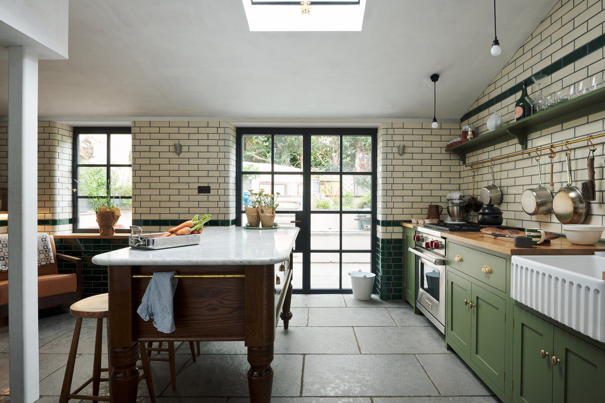 A quaint and modern Wilkes green Shaker kitchen features a cooker and sink run with a rustic oak worktop, a long shaker shelf, and a Dairy table with a breakfast nook. Adorned with a long hanging rail, sky light, glass doors, signature ceramics, brass cups, and a large wall of metro tiles it embodies a playful industrial and practical design.