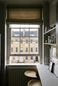 A stylish, mushroom-coloured Shaker galley kitchen features a cooker and sink run with Silestone worktop and a cosy breakfast nook. Beautifully decorated with simple, everyday kitchen items such as dried ingredients, muted ceramics, framed illustrations, deVOL Bum Chairs, a healing crystal, and an amazing view of Bath’s famous Georgian apartments, this calming and functional kitchen space blends character with tranquillity.