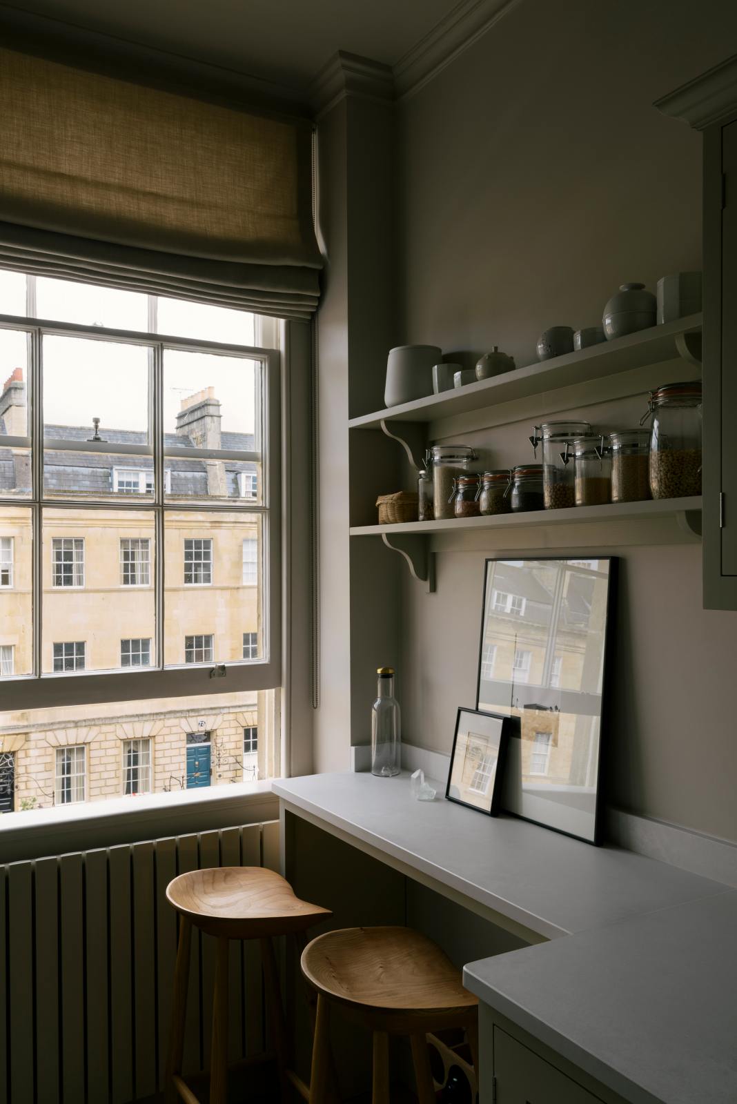 An angled-view of a stylish, mushroom-coloured Shaker galley kitchen features a cooker and sink run with Silestone worktop and a cosy breakfast nook. Beautifully decorated with simple, everyday kitchen items such as dried ingredients, muted ceramics, framed illustrations, deVOL Bum Chairs, a healing crystal, and an amazing view of Bath’s famous Georgian apartments, this calming and functional kitchen space blends character with tranquillity.