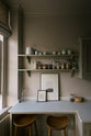 A stylish, mushroom-coloured Shaker galley kitchen features a cooker and sink run with Silestone worktop, wall cupboards and a cosy breakfast nook. Beautifully decorated with simple, everyday kitchen items such as dried ingredients, muted ceramics, framed illustrations, deVOL Bum Chairs, a healing crystal, and an amazing view of Bath’s famous Georgian apartments, this calming and functional kitchen space blends character with peace.