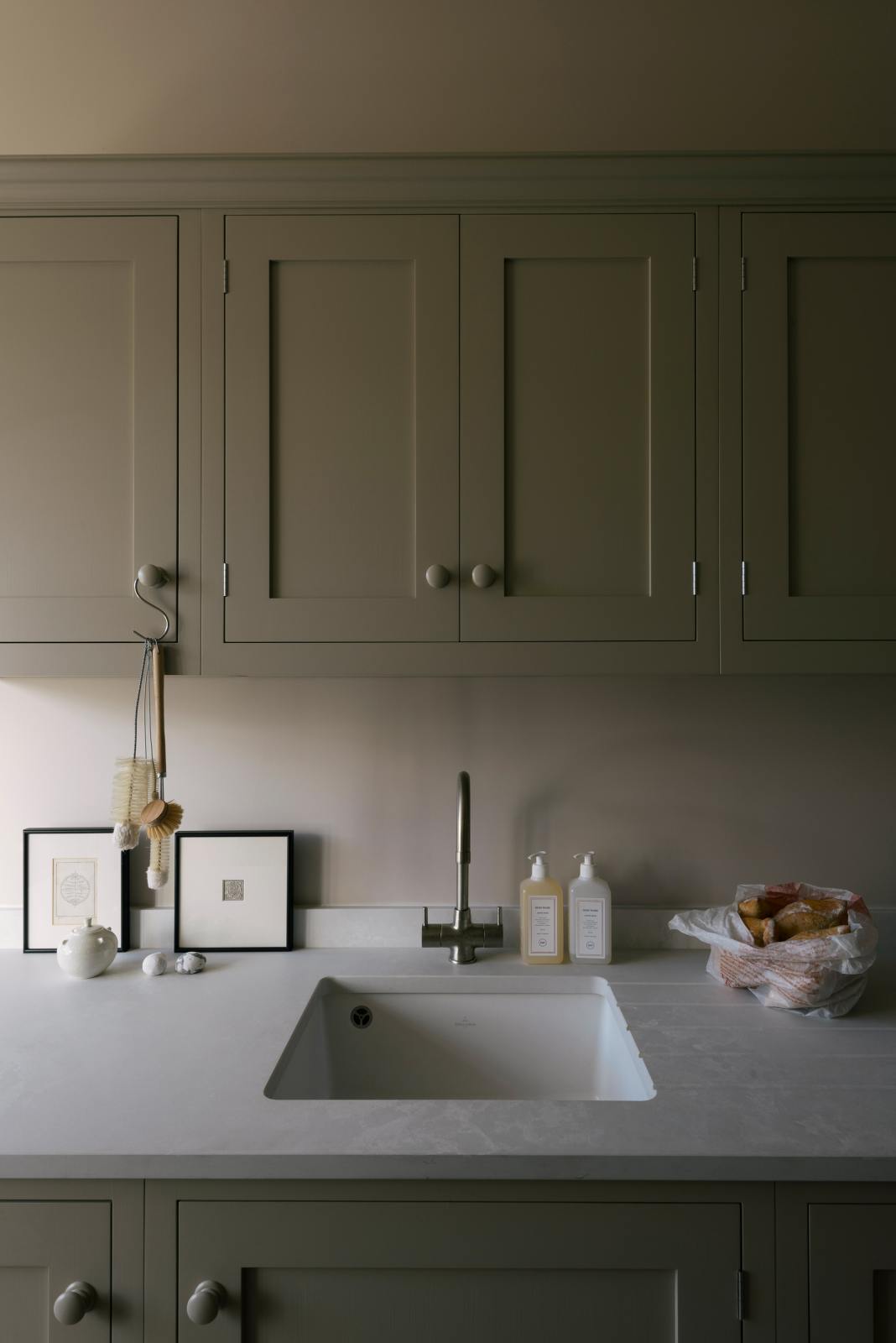 A stylish, mushroom-coloured Shaker galley kitchen features a cooker and sink run with Silestone worktop, wall cupboards and a single sink with a silver tap. Beautifully decorated with simple, everyday kitchen items such as muted ceramics, framed illustrations, a plastic bag full of bread, and small cleaning equipment, this calming and functional kitchen space blends character with peace.