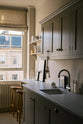An angled view of a stylish, mushroom-coloured Shaker galley kitchen features a cooker and sink run with Silestone worktop, wall cupboards and a cosy breakfast nook. Beautifully decorated with simple, everyday kitchen items such as dried ingredients, muted ceramics, framed illustrations, deVOL Bum Chairs, a healing crystal, and an amazing view of Bath’s famous Georgian apartments, this calming and functional kitchen space blends character with peace.