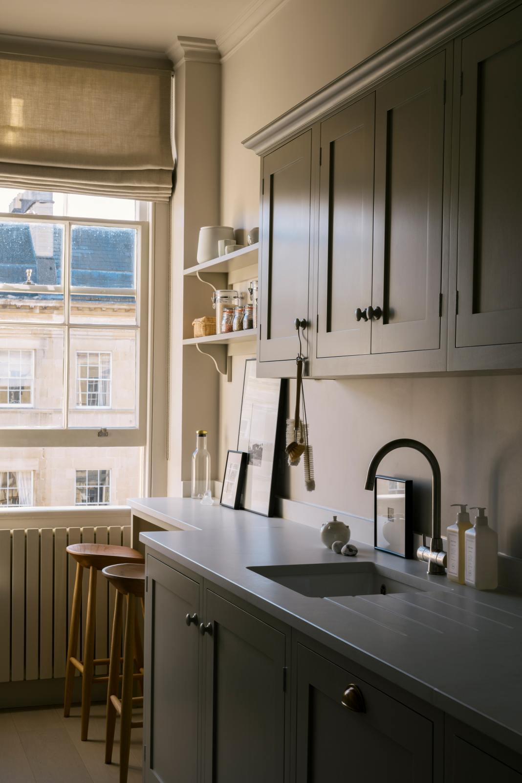 An angled view of a stylish, mushroom-coloured Shaker galley kitchen features a cooker and sink run with Silestone worktop, wall cupboards and a cosy breakfast nook. Beautifully decorated with simple, everyday kitchen items such as dried ingredients, muted ceramics, framed illustrations, deVOL Bum Chairs, a healing crystal, and an amazing view of Bath’s famous Georgian apartments, this calming and functional kitchen space blends character with peace.