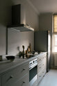 An angled view of a stylish, mushroom-coloured Shaker galley kitchen features a cooker and sink run with Silestone worktop. Beautifully decorated with simple, everyday kitchen items such as muted ceramics, wooden salt and pepper grinders, chopping boards, and an electric hob, this calming and functional kitchen space blends character with peace.