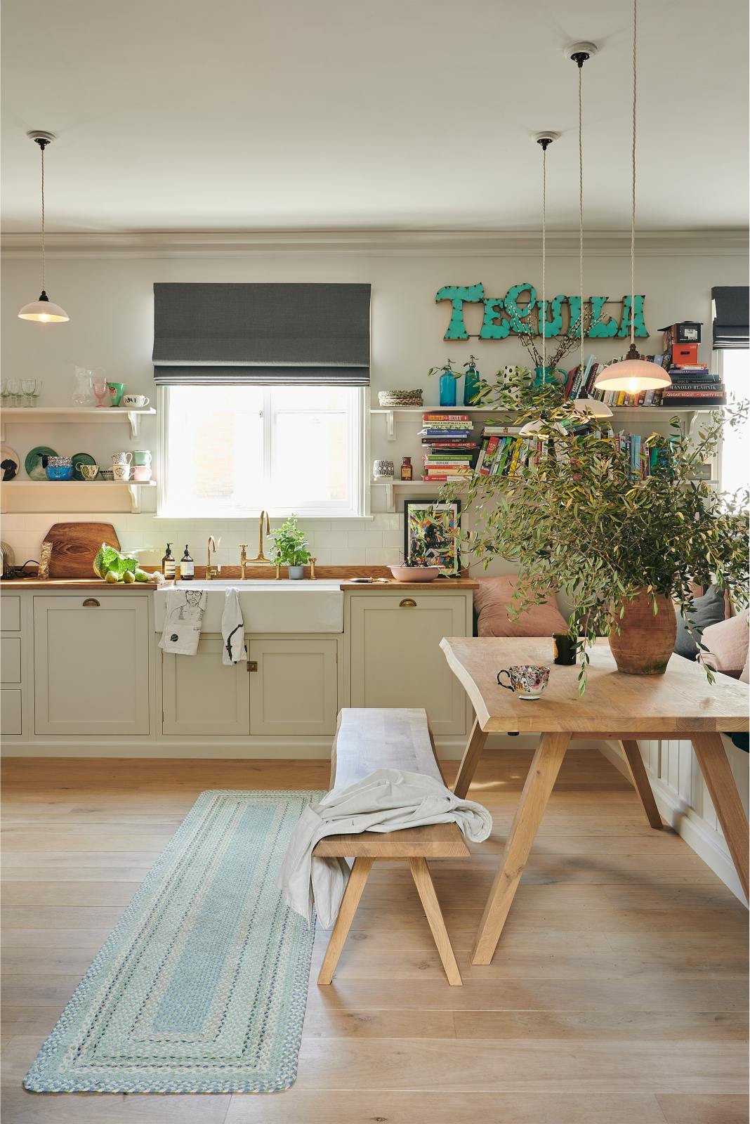 A vibrant Shaker kitchen features an L-shaped sink and cooker run with a solid oak worktop, a single Belfast sink, an aged brass Ionian tap, a Shaker shelf, and a bench. Lovingly decorated with a vintage “Tequila” sign, colourful cushions, overstocked books, a wild fauna arrangement, chattered skullcap porcelain lights, and a park bench dining table, it creates a characterful and inviting space.