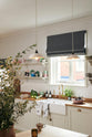 An oblique view of a vibrant Shaker kitchen features an L-shaped sink and cooker run with a solid oak worktop, a single Belfast sink, an aged brass Ionian tap, and Shaker shelves. Lovingly decorated with beautiful ceramics and drinking glasses, luscious green vegetables, and chattered skullcap porcelain lights, it creates a characterful and inviting space.