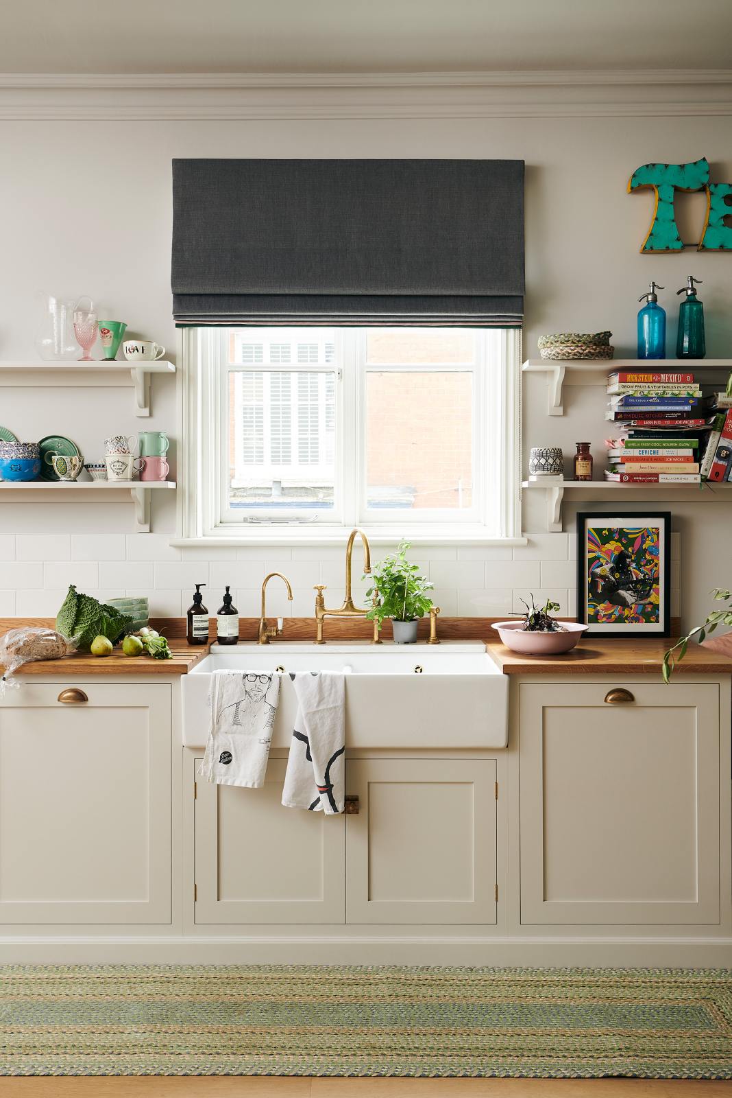 A vibrant Shaker kitchen features an L-shaped sink and cooker run with a solid oak worktop, a single Belfast sink, an aged brass Ionian tap, and Shaker shelves. Lovingly decorated with beautiful ceramics and drinking glasses, overstocked books, luscious green vegetables, chattered skullcap porcelain lights, and a vintage “Tequila” sign, it creates a characterful and inviting space.