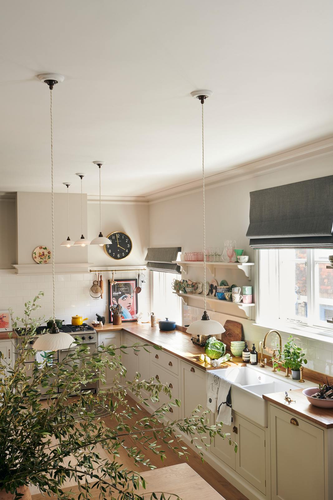 A vibrant Shaker kitchen features an L-shaped sink and cooker run with a solid oak worktop, a single Belfast sink, an aged brass Ionian tap, Shaker shelves, a silver gas cooker and white splash back tiles. Lovingly decorated with a gold-rimmed clock, an aged brass hanging rail, a chattered skullcap porcelain light, luscious green vegetables, beautiful ceramics, and a bold pop-art poster, it creates a characterful and inviting space.