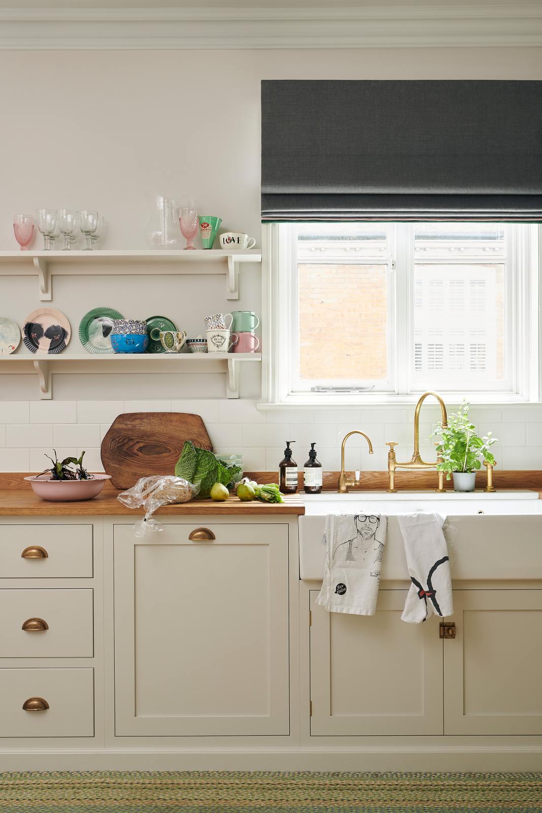 A vibrant Shaker kitchen features an L-shaped sink and cooker run with a solid oak worktop, a single Belfast sink, an aged brass Ionian tap, and Shaker shelves. Lovingly decorated with beautiful ceramics and drinking glasses, luscious green vegetables, and illustrative tea towels, it creates a characterful and inviting space.