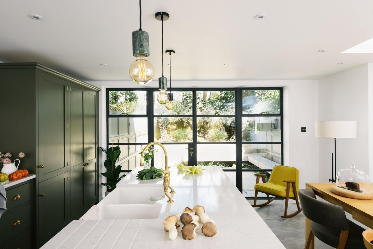 A bright and sophisticated Bakehouse Green shaker kitchen, styled with Scandinavian furniture and a soothing green palette. The kitchen features a cooker run, a central island with a double Belfast sink and Silestone worktop, and full-height cupboards. Natural light floods the kitchen and dining room through the glass French doors.