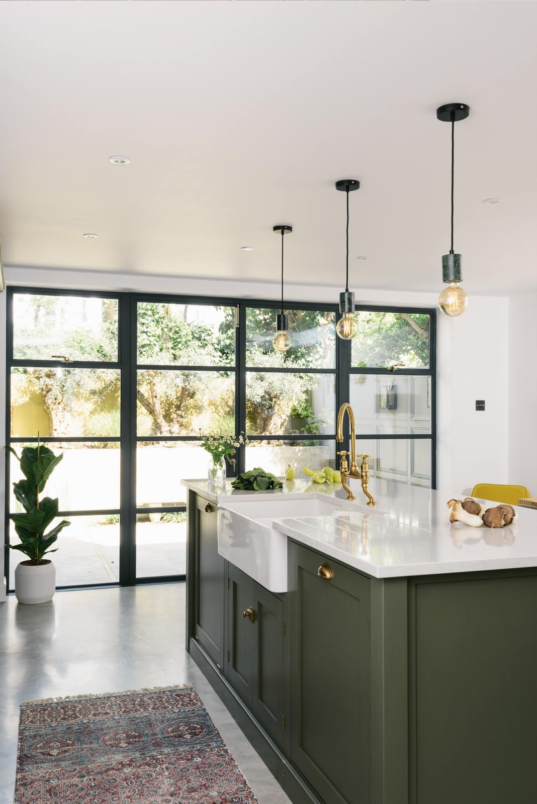 An angled view of a bright, sophisticated Bakehouse Green shaker kitchen, styled with Scandinavian furniture and a calming green palette. The kitchen showcases a central island with a double Belfast sink, Silestone worktop, and aged brass Ionian tap. Natural light floods the kitchen and dining room through the glass French doors.
