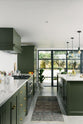 A bright and sophisticated Bakehouse Green shaker kitchen, styled with Scandinavian furniture and a soothing green palette. The kitchen features a cooker run, a central island with a double Belfast sink and Silestone worktop, and full-height cupboards. Natural light floods the kitchen and dining room through the glass French doors.