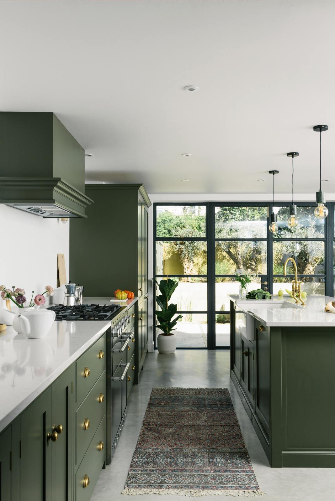 A bright and sophisticated Bakehouse Green shaker kitchen, styled with Scandinavian furniture and a soothing green palette. The kitchen features a cooker run, a central island with a double Belfast sink and Silestone worktop, and full-height cupboards. Natural light floods the kitchen and dining room through the glass French doors.