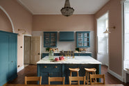 The room, a grand Georgian space from the 18th century, features charming pink walls. It showcases a modern Clerkenwell Blue shaker kitchen equipped with glazed wall cupboards, a shaker-style canopy, a cooker run, large full-height pantry cupboards, and a spacious kitchen island with a sink and an Arabescato marble worktop. Above is a beautiful vintage chandelier that enhances the renovated kitchen space and complements the esteemed property exceptionally well.