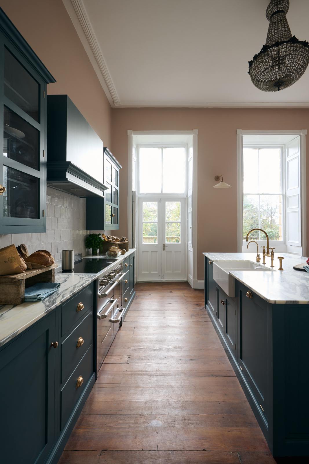 The room, a grand Georgian space from the 18th century, features charming pink walls. It showcases a modern Clerkenwell Blue shaker kitchen equipped with glazed wall cupboards, a shaker-style canopy, a cooker run, and a spacious kitchen island with a sink and an Arabescato marble worktop. The kitchen offers views of the balcony and garden through its original wooden floors and doors, and the renovated space complements the esteemed property beautifully.
