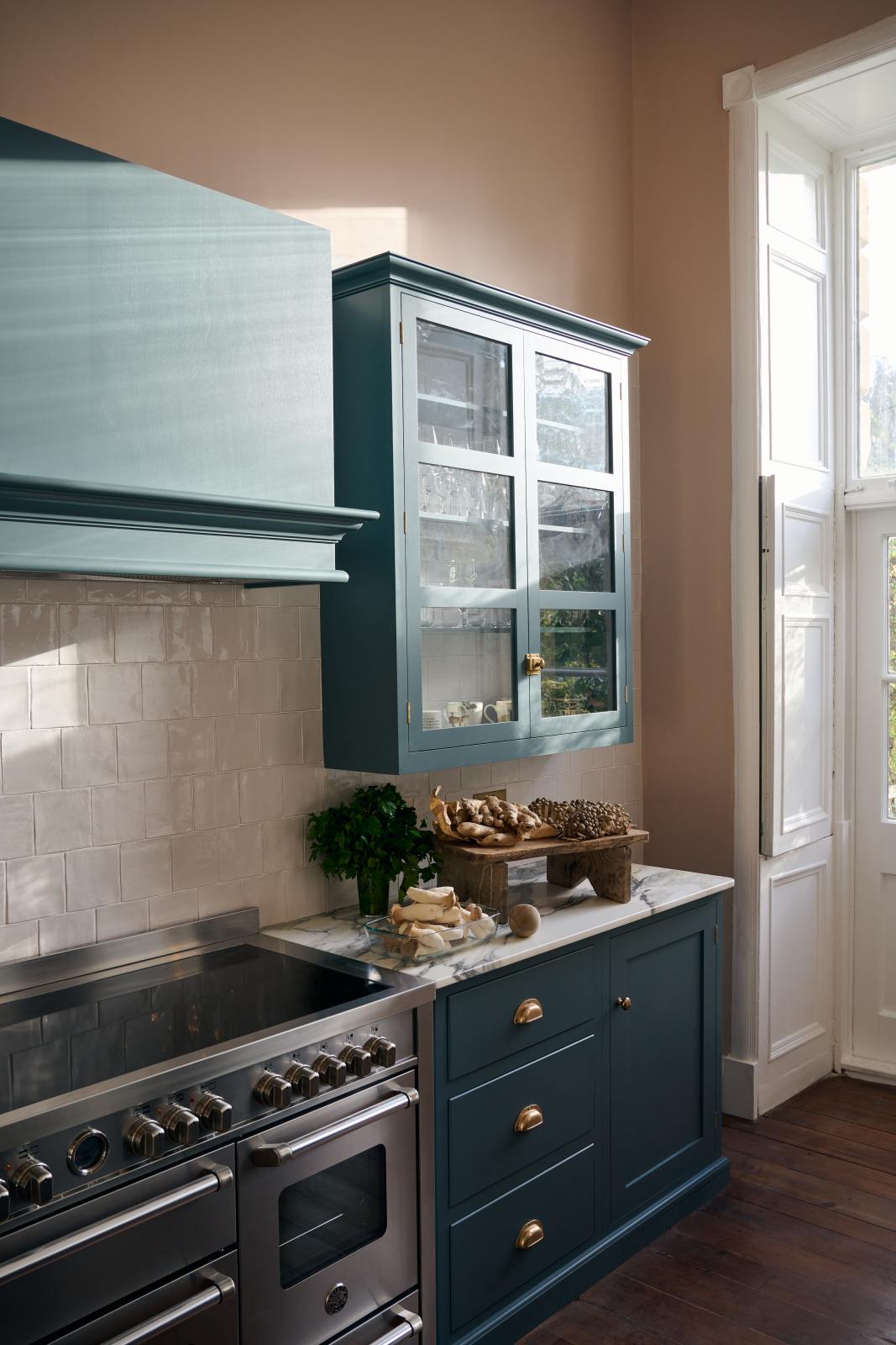 A closer look at a Clerkenwell Blue shaker kitchen’s cooker run equipped with Arabescato marble worktop, glazed wall cupboards, a shaker-style canopy and white porcelain splashback. Assorted fine drinking glasses and ceramic mugs are inside the glazed cupboard. On the worktop, there are herbs, bread, and fruits.
