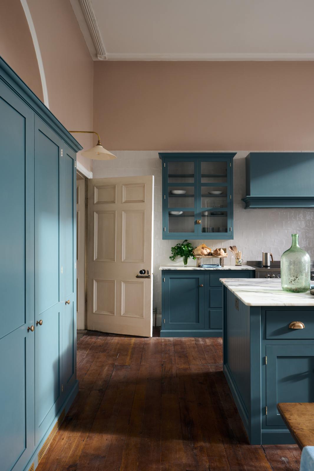 The room, a grand Georgian space from the 18th century, features charming pink walls. It showcases a modern Clerkenwell Blue shaker kitchen equipped with glazed wall cupboards, a shaker-style canopy, a cooker run, white porcelain splashback, large full-height pantry cupboards, and a spacious kitchen island with a sink and an Arabescato marble worktop.